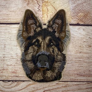 May include: Embroidered patch featuring a German Shepherd dog's face. The patch has a detailed design with brown, black, and gray thread, showcasing the dog's fur and facial features. The background is a light tan color.