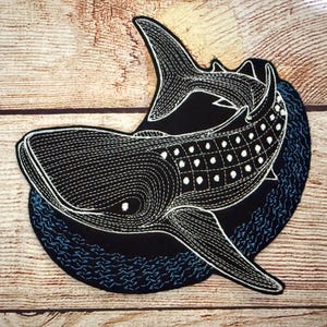 以下が含まれることがあります： 黒と白の刺繍パッチで、ジンベイザメのデザインです。ジンベイザメは白いステッチと白いドットパターンで詳細に描かれています。パッチは、青い波のようなディテールのある黒い背景に設定されています。