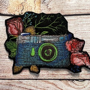Peut inclure: Patch brod&eacute; repr&eacute;sentant un appareil photo vintage avec un objectif vert entour&eacute; de fleurs et de feuillage. L'appareil photo est bleu et argent&eacute; sur un fond noir.