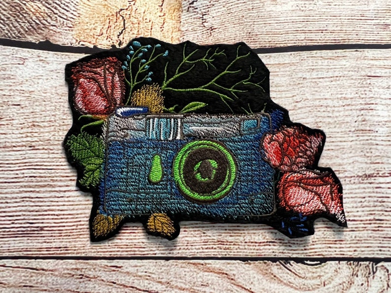 Peut inclure: Patch brod&eacute; repr&eacute;sentant un appareil photo vintage avec un objectif vert entour&eacute; de fleurs et de feuillage. L'appareil photo est bleu et argent&eacute; sur un fond noir.