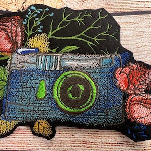 Peut inclure: Patch brod&eacute; d'un appareil photo bleu avec un objectif vert entour&eacute; de fleurs rouges et jaunes et de feuilles vertes sur un fond noir.