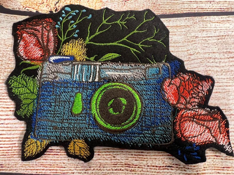 Peut inclure: Patch brod&eacute; d'un appareil photo bleu avec un objectif vert entour&eacute; de fleurs rouges et jaunes et de feuilles vertes sur un fond noir.