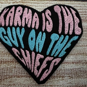 Puede incluir: Parche negro en forma de corazón con texto bordado en rosa y azul que dice "Karma is the guy on the sheets."