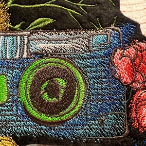 Peut inclure: Patch brod&eacute; d'un appareil photo bleu avec un objectif vert et une fleur rouge. L'appareil photo est entour&eacute; de feuilles vertes et d'une fleur jaune.