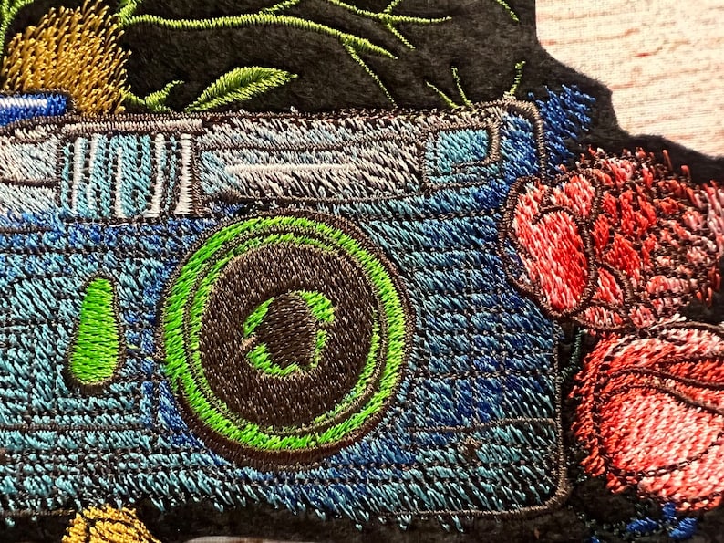 Peut inclure: Patch brod&eacute; d'un appareil photo bleu avec un objectif vert et une fleur rouge. L'appareil photo est entour&eacute; de feuilles vertes et d'une fleur jaune.