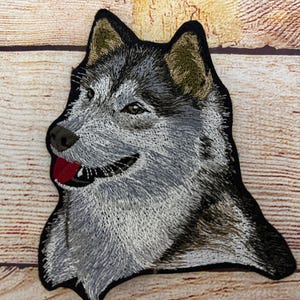 Puede incluir: Parche bordado con la cabeza detallada de un lobo en tonos grises, negros y beige. La boca del lobo está abierta, revelando una lengua roja. El parche tiene un borde negro y está sobre un fondo de madera.