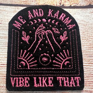 Może przedstawiać: Czarna, haftowana naszywka z różowym tekstem i wzorem. Na naszywce u góry widnieje napis "ME AND KARMA", a na dole "VIBE LIKE THAT". Wzór zawiera dłonie, księżyce, gwiazdy i promienie słoneczne.