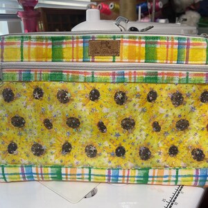 May include: A yellow and green plaid fabric zippered pouch with a sunflower and star print. The pouch has two zippered compartments.