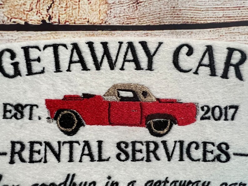 Puede incluir: Un letrero blanco con texto bordado en negro que dice "Getaway Car Est. 2017 - Rental Services -" con una ilustraci&oacute;n de un coche rojo convertible.