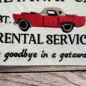 Puede incluir: Tela blanca bordada con el texto "Get Away Car" y "EST. 201..." y "RENTAL SERVICES" con una ilustraci&oacute;n de un coche vintage rojo y negro. El texto debajo del coche dice "Say goodbye in a getaway car."