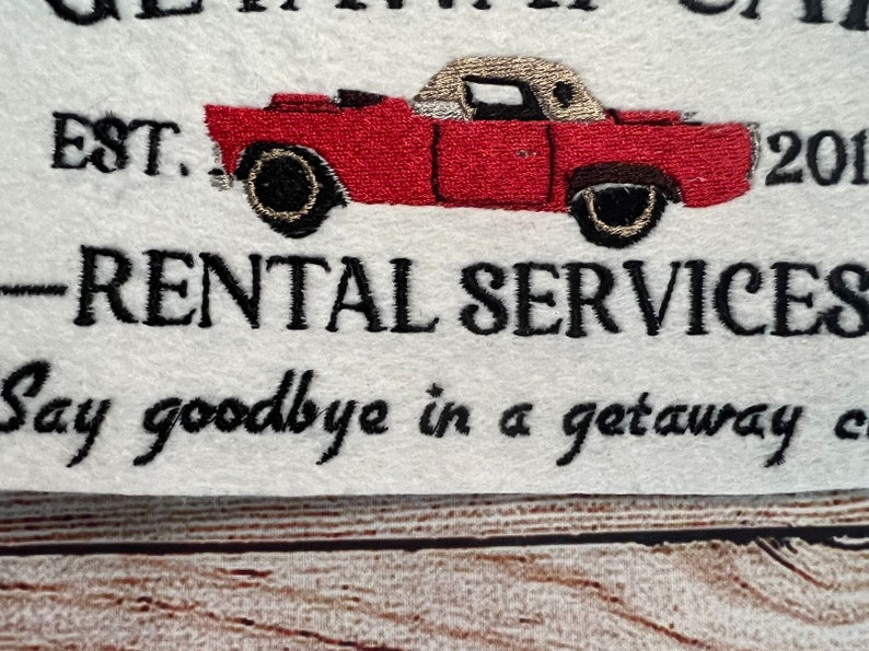 Puede incluir: Tela blanca bordada con el texto "Get Away Car" y "EST. 201..." y "RENTAL SERVICES" con una ilustraci&oacute;n de un coche vintage rojo y negro. El texto debajo del coche dice "Say goodbye in a getaway car."