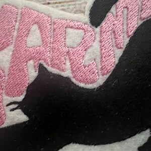 May include: Embroidered patch with the word "WARNING" in pink thread on a white background, and a black cat silhouette. Suitable for application on clothing or accessories.