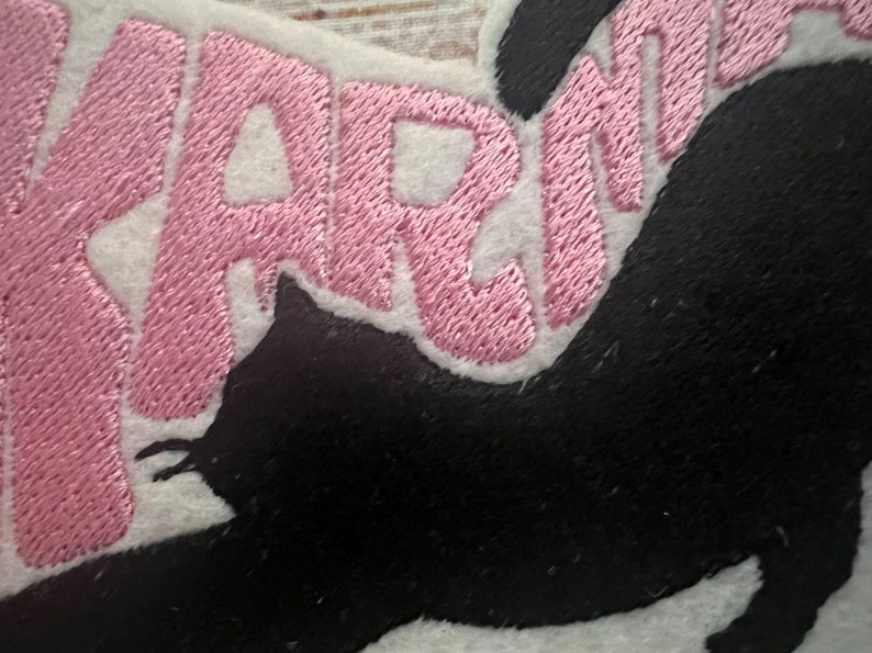 May include: Embroidered patch with the word "WARNING" in pink thread on a white background, and a black cat silhouette. Suitable for application on clothing or accessories.