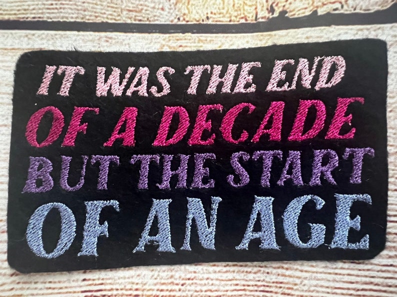 Puede incluir: Parche bordado negro con el texto "It was the end of a decade but the start of an age" en hilo rosa, morado y azul.