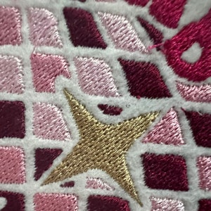 Pu&ograve; includere: Primo piano di una stella ricamata in oro su uno sfondo di tessuto bianco. La stella &egrave; circondata da quadrati rosa e bordeaux.