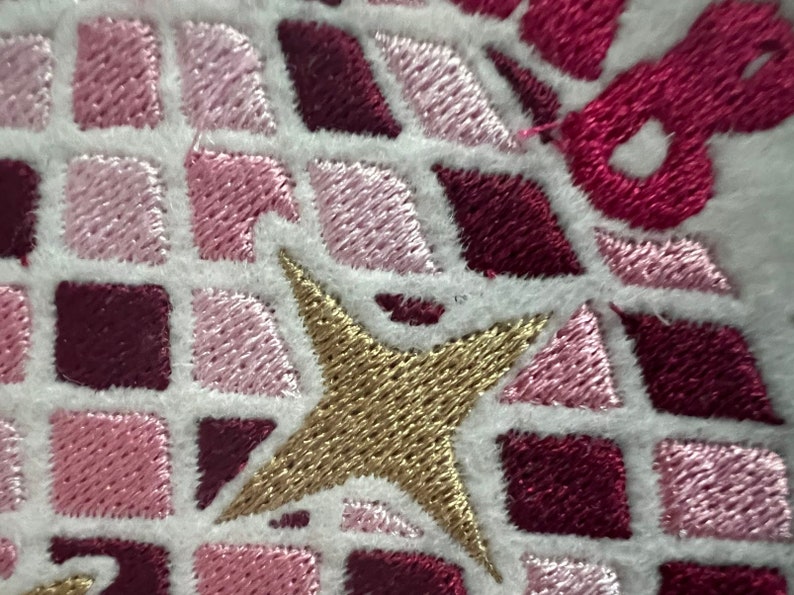 Pu&ograve; includere: Primo piano di una stella ricamata in oro su uno sfondo di tessuto bianco. La stella &egrave; circondata da quadrati rosa e bordeaux.