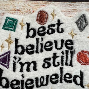 Puede incluir: Parche de tela blanca con texto bordado en negro que dice "best believe i'm still bejeweled". El parche est&aacute; decorado con piedras preciosas bordadas de colores y purpurina dorada.