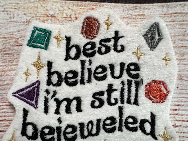 Puede incluir: Parche de tela blanca con texto bordado en negro que dice "best believe i'm still bejeweled". El parche est&aacute; decorado con piedras preciosas bordadas de colores y purpurina dorada.