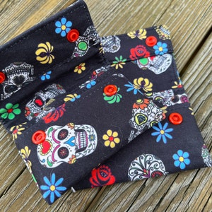 May include: A black fabric wallet with a colorful sugar skull and floral pattern. The wallet has red snap buttons and folds in half.