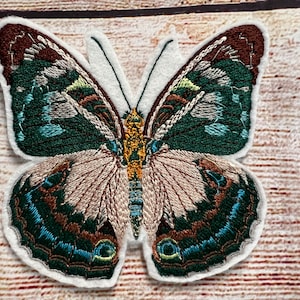 May include: Embroidered butterfly patch with intricate details. The wings feature shades of green, brown, and teal, with a cream-colored body and white border. This decorative patch is ideal for adding flair to clothing or accessories.