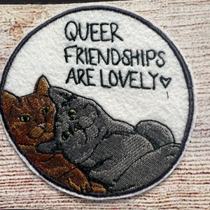 May include: Embroidered patch with two cats cuddling, one brown and one gray. The text reads "Queer Friendships Are Lovely" with a small heart.
