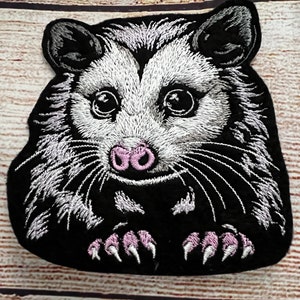 May include: Embroidered patch of a possum with a black and white fur pattern. The possum has a pink nose and is looking directly at the viewer. The patch is on a black background.