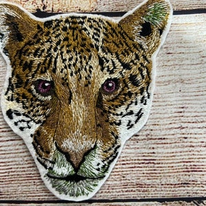 May include: Embroidered patch of a jaguar's face with brown, black, and white fur. The jaguar has pink eyes and a green nose.