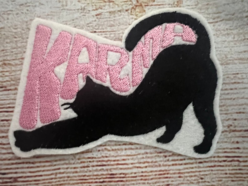 May include: Embroidered patch with a black cat stretching, and the word "KARMA" in pink. The patch has a white border. It is designed to be ironed or sewn onto fabric, suitable for clothing or accessories.