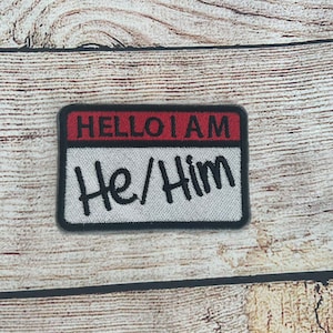 Può includere: Un'etichetta per il nome in bianco e nero con bordo rosso e nero. L'etichetta recita "HELLO I AM He/Him".