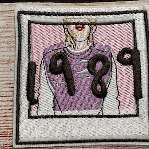 Peut inclure: Un patch brodé blanc et noir avec un design de cadre photo. La photo à l'intérieur montre une personne portant un pull violet avec le numéro "1989" dessus. La personne a les cheveux blonds et porte du rouge à lèvres rouge.