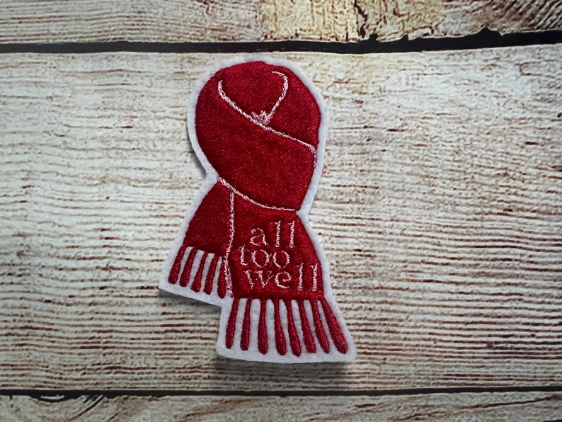 Puede incluir: Parche bordado rojo con un contorno blanco de una bufanda y el texto "all too well" en blanco.