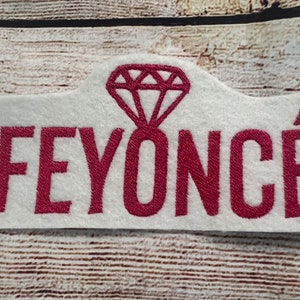 Puede incluir: Parche de fieltro blanco con la palabra "FEYONCÉ" bordada en burdeos con un diamante burdeos encima.
