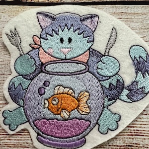 Könnte beinhalten: Gestickter Aufnäher mit einer Cartoon-Katze, die eine Gabel und ein Messer hält und vor einem Fischglas mit einem orangefarbenen Fisch sitzt. Die Katze ist blau und lila, mit einer rosa Schleife. Das Fischglas ist lila und der Fisch ist orange.