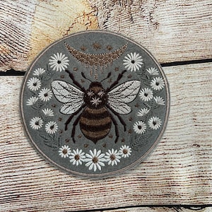 Może przedstawiać: Szara naszywka haftowana z pszczołą w środku, otoczona białymi kwiatami i rosnącym księżycem z gwiazdami nad nią. Pszczoła ma brązowo-czarne paski i białe skrzydła.