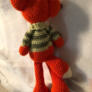 May include: A crocheted orange fox plush toy with a green and beige striped sweater. The fox is standing on a white surface.