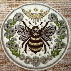 Puede incluir: Un parche blanco bordado con una abeille en el centro, rodeada de flores y una luna creciente con estrellas. La abeille es marrón y amarilla con un fondo blanco. Las flores son grises y verdes.