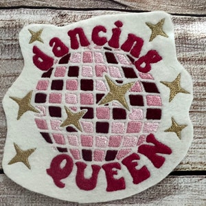 Può includere: Un cerotto di feltro bianco con una palla da discoteca rosa e bordeaux, stelle dorate e il testo "dancing queen" in rosa.