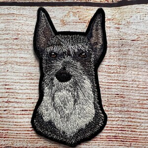 Puede incluir: Parche bordado de la cabeza de un perro schnauzer negro y gris. El perro tiene barba y bigote blancos.