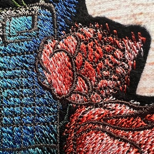 Peut inclure: Gros plan sur un patch brod&eacute; avec un motif floral. Le patch pr&eacute;sente une fleur rouge, blanche et bleue avec une tige verte. Le patch est fait d'un tissu doux et textur&eacute;.