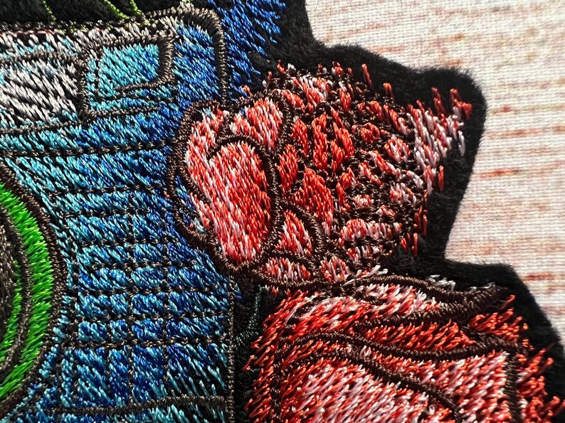 Peut inclure: Gros plan sur un patch brod&eacute; avec un motif floral. Le patch pr&eacute;sente une fleur rouge, blanche et bleue avec une tige verte. Le patch est fait d'un tissu doux et textur&eacute;.