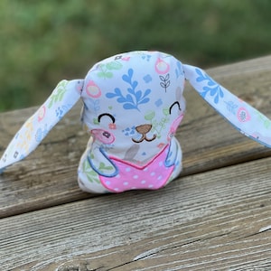 May include: A plush bunny toy with a white background and floral pattern. The bunny has pink and white polka dot fabric on its chest and embroidered eyes and nose.