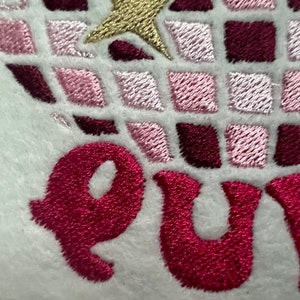 Pu&ograve; includere: Toppa ricamata con un design geometrico rosa e bordeaux e la parola "QUE" in filo rosa.