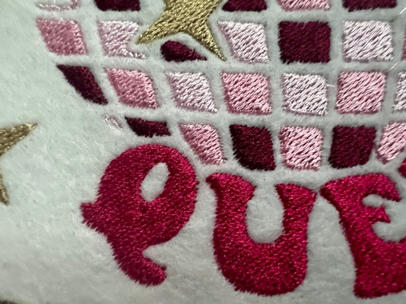 Pu&ograve; includere: Toppa ricamata con un design geometrico rosa e bordeaux e la parola "QUE" in filo rosa.