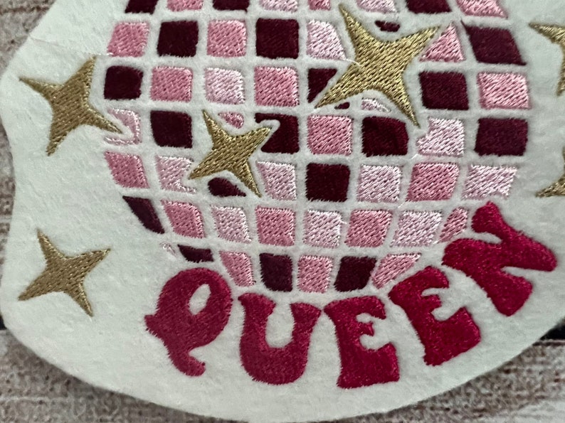 Pu&ograve; includere: Toppa ricamata da stirare con un design a palla da discoteca rosa e bordeaux con stelle dorate e la parola "QUEEN" in rosa.