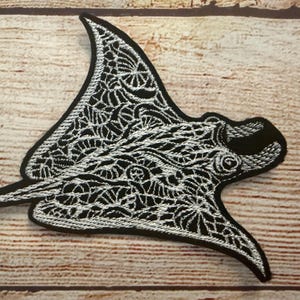 May include: Black and white embroidered patch in the shape of a manta ray. The patch features intricate white stitching on a black background, with a detailed design. The patch is placed on a wooden surface.