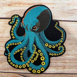 Puede incluir: Un parche bordado que presenta un pulpo de color verde azulado con detalles en marrón oscuro en la cabeza y ventosas circulares de color amarillo brillante a lo largo de sus tentáculos. El pulpo está sobre un fondo negro con costuras detalladas.