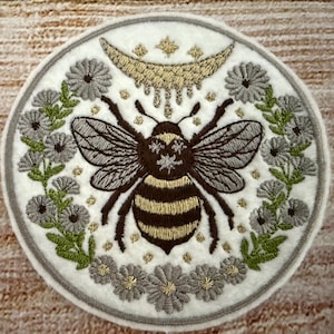 May include: A white embroidered patch with a bee in the center, surrounded by flowers and a crescent moon with stars. The bee is brown and yellow with a white background. The flowers are gray and green.