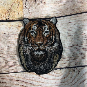 May include: Embroidered tiger patch showing a detailed tiger face with brown, black, and white thread. The patch has a black border and is designed to be sewn or ironed onto fabric. Suitable for jackets, bags, or other apparel.