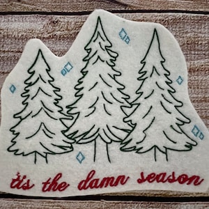 May include: White felt patch with an embroidered design of three green pine trees and blue snowflakes. The text "it's the damn season" is embroidered in red at the bottom.