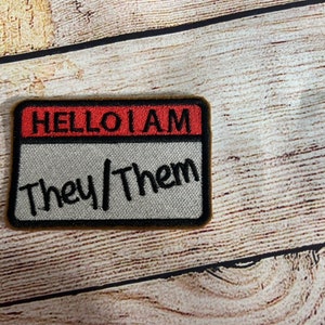 Puede incluir: Un parche bordado marrón y negro con una etiqueta de nombre roja y blanca que dice "Hello I am They/Them".